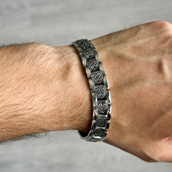 Ett brett silverfärgat länkarmband i metall på en mans handled.